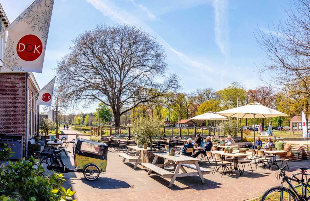 Het terras van De Oude Keuken op een zonnige voorjaarsdag, met gasten aan de picknicktafels onder de parasols en de DOK-vlaggen aan de gevel.