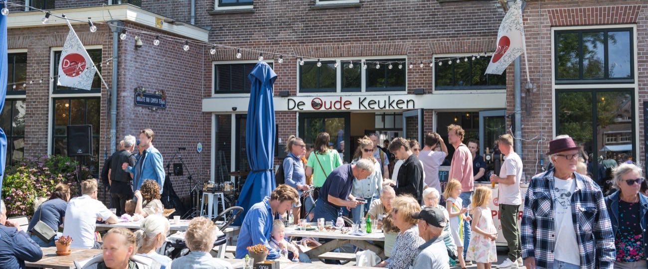 Gasten aan de picknicktafels op het volle terras van De Oude Keuken in Castricum tijdens het zomerfestival, met lichtslingers en DOK-vlaggen aan de bakstenen gevel en de naam boven de ingang.
