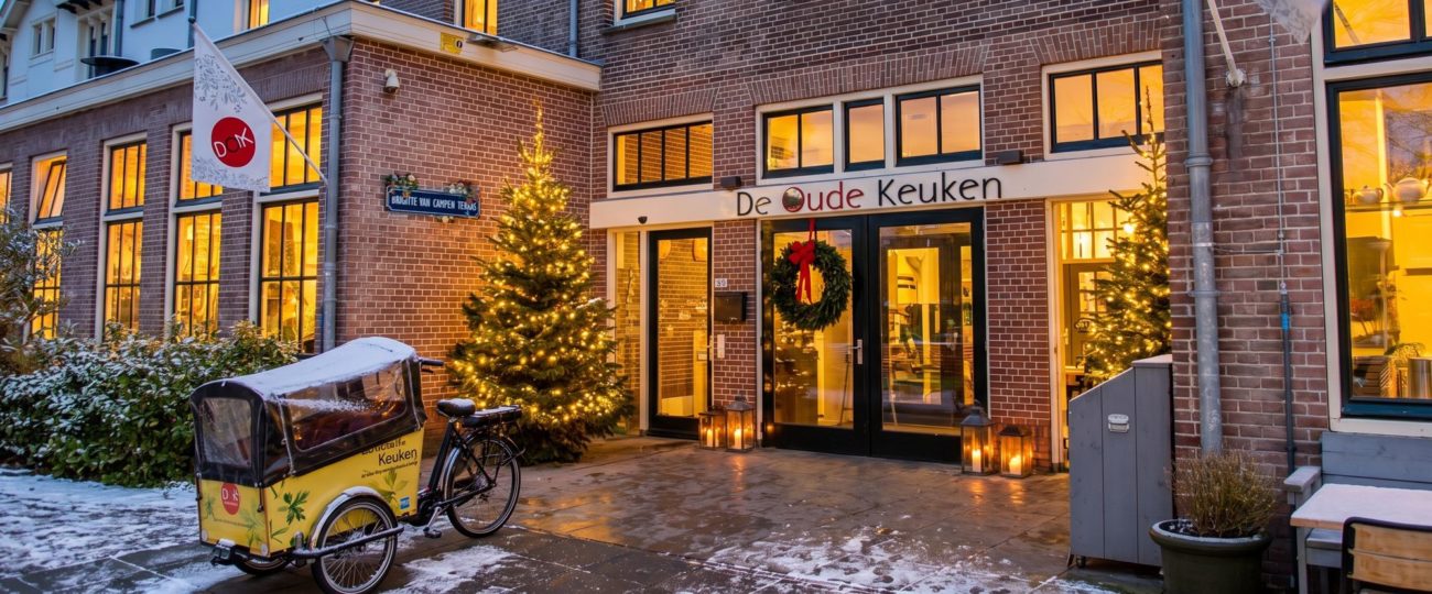 De gevel van De Oude Keuken in Castricum bij avond in kerstsfeer, met verlichte kerstbomen, een krans op de deur en lantaarns bij de ingang