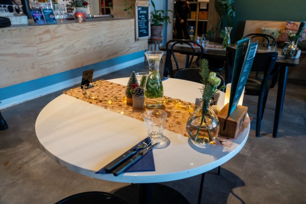 Een gedekte ronde tafel in De Oude Keuken met een gouden tafelloper, dennenappels, een klein kerstboompje en een karaf water, met kerst 2025